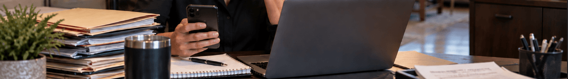 Person holding a smartphone at a busy desk with a laptop and stacks of papers.